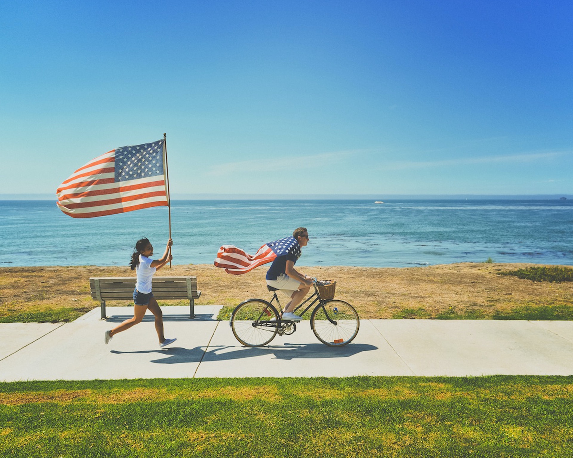 Labor Day 2023: Celebrating the American Workforce and Its Achievements by young woman carrying the American flag running behind a young man riding a bike and carrying the American flag on his back 