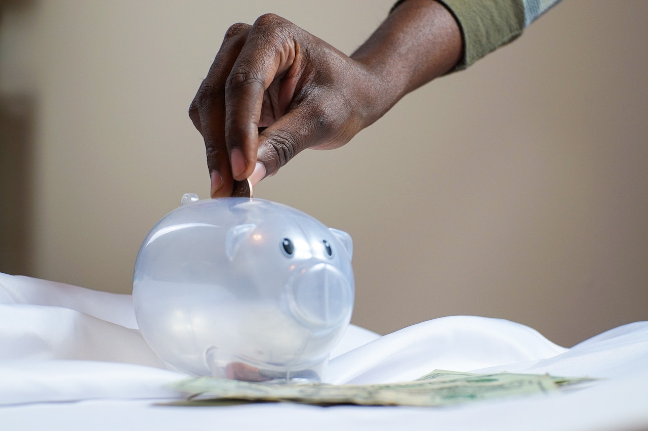 Black Person's Hand Putting Coin in a Piggy Bank that is placed on sheets with few dollar bills on top of it